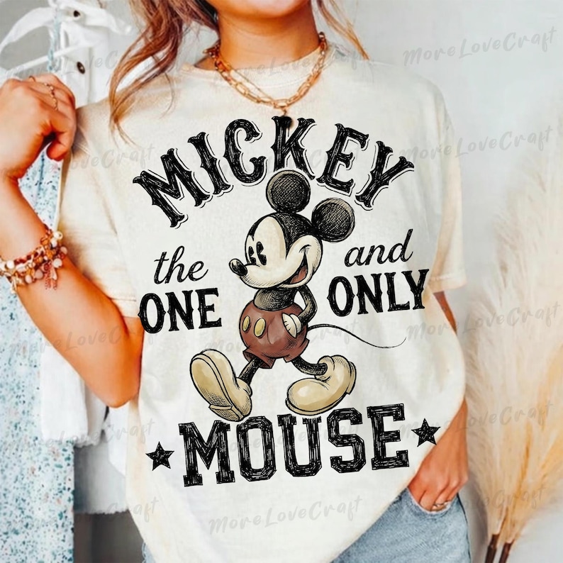 May include: Cream-colored t-shirt featuring a graphic of Mickey Mouse with the text "MICKEY the ONE and ONLY MOUSE" in black. Mickey is depicted in his classic attire, with brown shorts and yellow shoes. The design includes black stars.