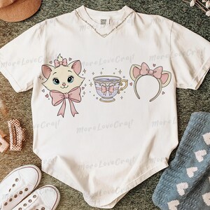 May include: White t-shirt featuring a cartoon cat face, teacup, and headband with cat ears, all in pink and light purple. The cat has a pink bow and the teacup has a bow. The shirt has the text "More Love Craft".