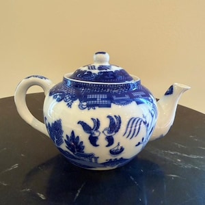 Puede incluir: Una tetera de cerámica blanca y azul con tapa. La tetera presenta un diseño clásico azul y blanco de patrón Willow. El asa y el pico son blancos. La tetera es de tamaño tradicional.