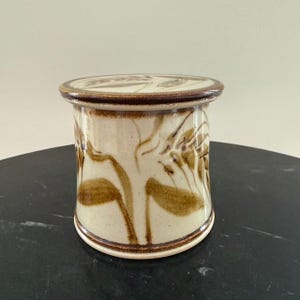 Puede incluir: Un tarro de cerámica con tapa, con una base de color crema y diseños florales marrones. El tarro tiene forma cilíndrica con una tapa redondeada y un borde marrón oscuro. El patrón floral se repite en la tapa.