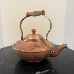 Op de afbeelding: Een koperen theeketel met een brede, ronde behuizing, een korte gebogen tuit en een afneembaar deksel met een kleine knop. De ketel is voorzien van een hoog, gebogen messing handvat met een gladde, cilindervormige houten greep.