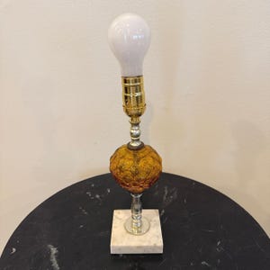 May include: A vintage table lamp with a white bulb, brass socket, and a textured amber glass globe. The lamp is supported by a silver stem and a square marble base. The lamp is approximately 12 inches tall.