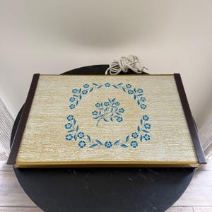 Puede incluir: Bandeja rectangular con diseño floral. La bandeja tiene una superficie beige claro con una corona floral azul y una ramita floral central. Tiene un borde marrón oscuro y un cable de alimentación.