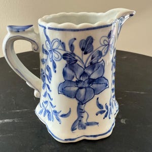 May include: White ceramic pitcher with a blue floral design. The pitcher has a curved handle and a spout for pouring. The floral design includes a large flower and leaves. The rim of the pitcher has a scalloped edge.