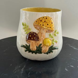 May include: Cream-colored ceramic vase with a textured surface and a yellow interior. The vase features a raised relief design of three mushrooms in shades of brown and yellow, with green leaves and grass.
