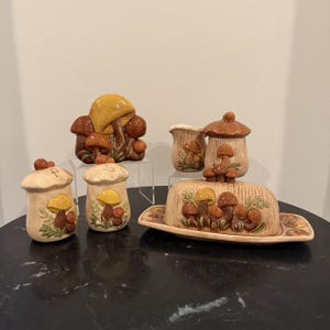 Puede incluir: Un juego de cocina de cerámica que incluye una mantequera, una jarra de leche y saleros y pimenteros, decorados con motivos de setas en relieve pintados a mano en marrón, amarillo y naranja sobre un fondo texturizado de color beige claro.