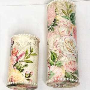 Pode incluir: Duas velas de pilar decorativas com desenhos florais. As velas apresentam um fundo creme com rosas rosa e amarelas, folhas verdes e uma borda de contas de pérolas na parte superior e inferior. Uma vela é mais alta que a outra.