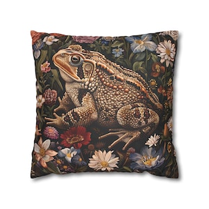May include: A square throw pillow with a dark background featuring a detailed illustration of a brown toad sitting on a bed of colorful flowers.