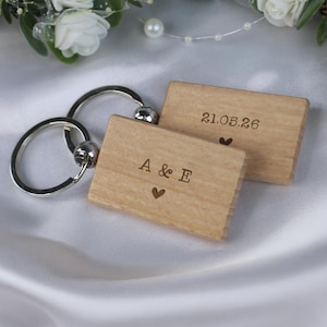 Puede incluir: Dos llaveros de madera con anillas plateadas. Un llavero tiene las letras "A & E" y un corazón grabado. El otro llavero tiene la fecha "21.05.26" y un corazón grabado. Los llaveros son de color marrón claro.