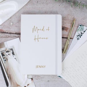 May include: White notebook with a gold inscription that reads "Maid of Honour" and the name "JENNY".