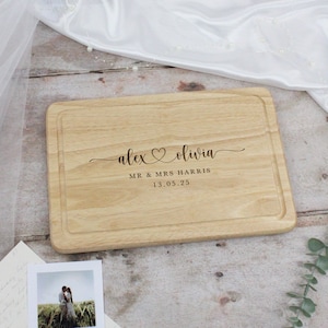 Könnte beinhalten: Personalisiertes Holz-Schneidebrett mit den Namen "alex" und "olivia", die in Kursivschrift mit einem Herzsymbol zwischen den Namen graviert sind. Der Text "MR & MRS HARRIS" und "13.05.25" ist ebenfalls auf das Brett graviert.