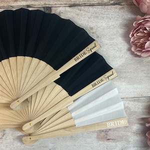 Peut inclure: Éventails pliants avec des manches en bois et du tissu noir ou blanc. Les éventails portent les inscriptions "BRIDE Squad" et "BRIDE". Des fleurs roses sont en arrière-plan. Ces éventails sont parfaits pour une fête de mariage.