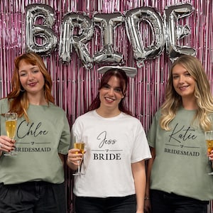 May include: Two sage green t-shirts and one white t-shirt featuring black printed text. The sage shirts display the names Chloe and Katie above the word BRIDESMAID. The white shirt displays the name Jess above the word BRIDE.