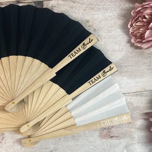 May include: Wooden hand fans with black and white pleated fabric. The fans have the phrases "TEAM Bride" and "THE Bride" printed on the wooden handles. Pink flowers are in the background.
