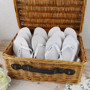 May include: A woven wicker basket containing several white flip-flops. Each flip-flop features white straps and the text "L&K" or "16.08.25". The basket has a dark brown handle and is open, displaying the contents.