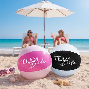May include: Two beach balls with "TEAM Bride" text, one pink and white, the other black and white, on a sandy beach. Heart-shaped sunglasses and a starfish are on the sand. Two people relax in the background under a parasol.