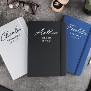 May include: Three notebooks with different colored covers and personalized text. The covers are white, black, and blue. The text on the covers reads "Charlie,  Groomsman 18.07.25", "Arthur, Groom 18.07.25", and "Freddie, Best Man 18.07.25".