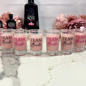 May include: A collection of shot glasses filled with a pink liquid, each bearing the text "TEAM Bride" in pink and black script. The glasses are arranged in a row, with bottles of Tequila Rose in the background, suggesting a bridal party theme.