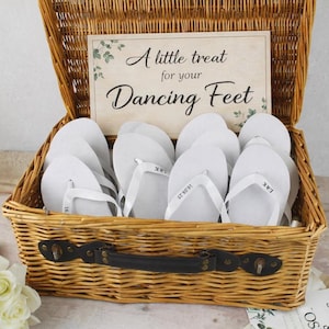 May include: A woven wicker basket containing white flip-flops with white straps. A wooden sign inside the basket reads "A little treat for your Dancing Feet". The flip-flops have the initials "L&K" and the date "16.08.25" printed on them.