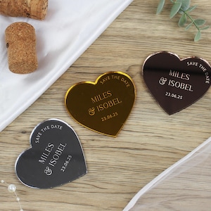 May include: Three gold, silver, and rose gold heart-shaped save the date cards with the text "Save the Date" and "Miles & Isobel" with the date "23.06.25" printed on each card.