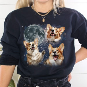 Może przedstawiać: Granatowa bluza z nadrukiem przedstawiającym trzy psy rasy corgi i pełnię księżyca. Corgi mają ekspresyjne twarze na tle gwiezdnego nieba. Unikalny i zabawny design dla miłośników psów.
