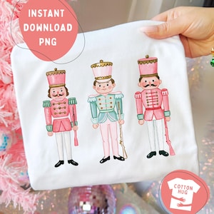 May include: A white t-shirt featuring a graphic illustration of three nutcracker figures. The nutcrackers are dressed in pink and mint green uniforms with gold accents, each holding a rifle. The design is centered on the front of the garment.