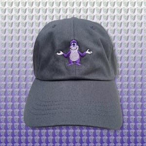 Pode incluir: Um boné de beisebol cinza escuro com um personagem de desenho animado roxo bordado com os braços estendidos. O personagem tem um rosto branco e um corpo roxo.