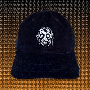 Pode incluir: Boné de beisebol de veludo cotelê preto com um gráfico bordado branco de um rosto estilizado. O boné tem uma aba curva e um design de seis painéis. O fundo é um padrão repetido de um rosto semelhante.