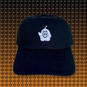 Pode incluir: Boné de beisebol preto com um design de fantasma branco bordado. O fantasma tem um rosto sorridente e está acenando. O boné tem uma aba curva e uma alça ajustável na parte de trás.