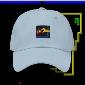 Pode incluir: Um boné de beisebol azul claro com uma imagem bordada de uma cobra comendo uma maçã. A imagem está em uma moldura preta com uma borda azul.