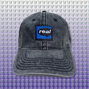 Könnte beinhalten: Eine schwarze Baseballkappe aus Denim mit einem blauen und schwarzen quadratischen Patch, auf dem in weißer Schrift "real" steht.