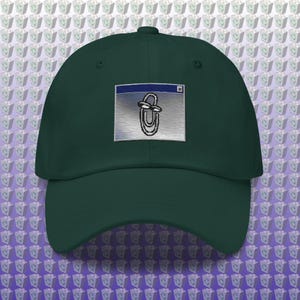 May include: A dark forest green baseball cap featuring an embroidered patch on the front. The patch displays a classic computer interface window containing a stylized paperclip character with eyes, rendered in black, white, and blue tones.