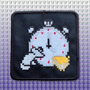 May include: A black embroidered patch with a white pixelated image of a clock with a red cross inside. The clock is surrounded by red dots and a yellow pixelated object.