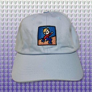 May include: A light blue baseball cap with an embroidered pixelated image of a person with blonde hair wearing a red shirt and blue pants.