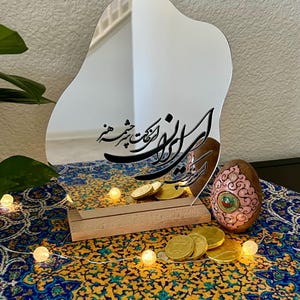 May include: A decorative mirror with an irregular shape, featuring calligraphy in black. The mirror is propped up on a wooden base, with gold coins and a decorated egg nearby. The background includes a patterned cloth and string lights.