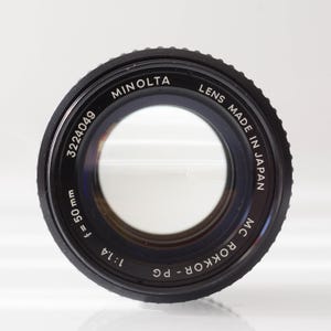 Op de afbeelding: Een zwarte Minolta MC Rokkor-PG cameralens met de tekst "LENS MADE IN JAPAN". De lens heeft een brandpuntsafstand van 50 mm en een diafragma van f/1.4. Het serienummer is 3224049.