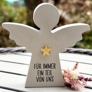 Könnte beinhalten: Graue Engelsfigur mit ausgebreiteten Flügeln und einem goldenen Stern auf der Brust. Der Text "FÜR IMMER EIN TEIL VON UNS" ist schwarz gedruckt. Ein kleiner Strauß Trockenblumen liegt neben dem Engel.
