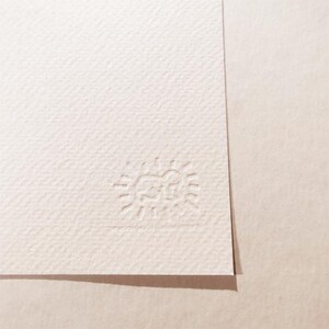 May include: A close-up of a white textured paper with a small embossed logo in the bottom right corner. The logo is a simple sun with a face inside.