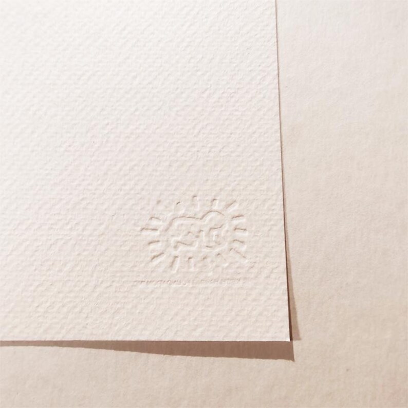 May include: A close-up of a white textured paper with a small embossed logo in the bottom right corner. The logo is a simple sun with a face inside.