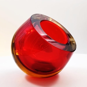 Puede incluir: Un cuenco de cristal esférico con un interior rojo vibrante y una capa exterior de cristal transparente grueso. El cuenco tiene una abertura circular inclinada en la parte superior, creando una silueta angular con bordes pulidos.