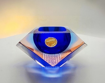 May include: A diamond-shaped art glass bowl with a deep blue interior and a gold-colored emblem. The bowl has a clear, angled exterior with hints of peach and blue. The base of the bowl has a textured, crystal-like design.