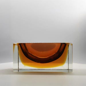 May include: A rectangular glass bowl featuring a clear outer frame and a deep amber-colored interior basin. The design consists of a thick, transparent glass exterior surrounding a curved, bowl-shaped center with a rich, warm orange-brown hue.