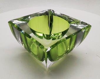 May include: A square green glass ashtray with a clear glass rim. The ashtray has a slightly rounded bottom and is made of thick glass.