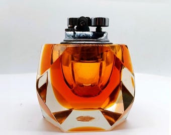 May include: A table lighter featuring a faceted, amber-colored glass base with a geometric design. The top mechanism is constructed from polished silver-toned metal, including a circular ignition button and a small adjustment dial for the flame.