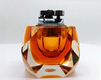 May include: A vintage table lighter with a faceted, orange-tinted glass body and a chrome-colored metal top. The lighter has a geometric design and is set against a white background. The lighter is a decorative item.