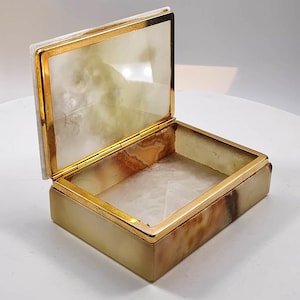 Könnte beinhalten: Eine rechteckige Scharnierbox aus poliertem Stein mit einer durchscheinenden, marmorierten Optik in Creme-, Beige- und Bernsteintönen. Die Box verfügt über einen metallischen, goldfarbenen Rahmen an den Kanten und ein passendes Scharnier am Deckel.