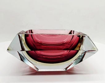 May include: A faceted, ruby-colored glass bowl with a square shape. The bowl has a deep, circular interior and clear, angled sides. The glass has a polished finish, reflecting light and creating a striking visual effect. A decorative piece.