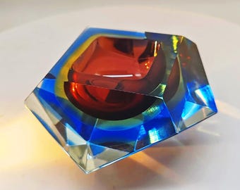 May include: A geometric ashtray with a clear, faceted exterior. The interior features a deep red bowl, surrounded by bands of blue and yellow. The ashtray is a decorative item.