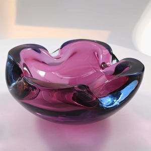 Puede incluir: Un cuenco de cristal de color púrpura intenso con un borde grueso, orgánico y ondulado. El material translúcido presenta tonos variados de violeta y magenta, creando una forma lisa, redondeada y asimétrica que refleja la luz en su superficie.