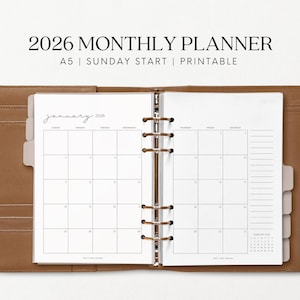 May include: Open tan leather A5 planner displaying a 2026 monthly calendar. The calendar is open to January, with a simple layout and Sunday start. The planner includes tabs and a calendar for February 2026. Text at the top reads "2026 MONTHLY PLANNER".